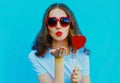 Portrait of beautiful young woman blowing red lips sweet air kiss with red heart shaped lollipop wearing sunglasses