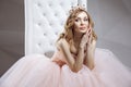 Portrait of beautiful young woman in crown and luxurious pink dress posing on stylish white armchair. sitting, touching her face Royalty Free Stock Photo