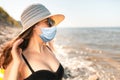 Portrait of a beautiful young woman, with full Breasts, in a bathing suit, wearing a hat and a medical mask. Beach and sea, windy Royalty Free Stock Photo