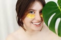 Portrait of beautiful young woman with golden eye patches at green monstera leaf on white. Happy girl with natural skin and Royalty Free Stock Photo