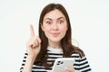 Portrait of beautiful young woman, has an idea, suggestion, shows one finger and holds smartphone, points up, white Royalty Free Stock Photo