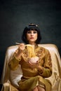 Portrait of beautiful young woman in image of egyptian queen, Cleopatra eating instant noodles against dark vintage