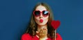 Portrait of beautiful young woman with lollipop blowing red lips sending sweet air kiss wearing a red heart shaped sunglasses Royalty Free Stock Photo