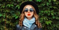 Portrait of beautiful young woman model wearing a black round hat, jacket, sunglasses over green leaves wall Royalty Free Stock Photo