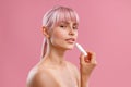 Portrait of beautiful young woman with naked shoulders holding lip balm near her lips isolated over pink background Royalty Free Stock Photo