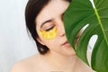 Portrait of beautiful young woman with natural makeup and golden eye patches at green monstera leaf on white background. Natural Royalty Free Stock Photo