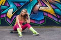 Portrait of beautiful young woman in pink bodysuit sitting on skateboard near wall with graffiti