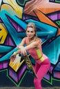 Portrait of beautiful young woman in pink bodysuit with skateboard posing near wall with graffiti