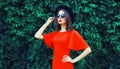 Portrait of beautiful young woman posing in red dress, black round hat on green leaves wall background Royalty Free Stock Photo