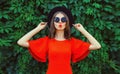 Portrait of beautiful young woman posing in red dress, black round hat on green leaves wall background Royalty Free Stock Photo