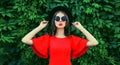 Portrait of beautiful young woman posing in red dress, black round hat on green leaves wall background Royalty Free Stock Photo