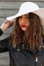 Portrait beautiful young woman with sexy red lips in black fashion leather jacket in white trendy summer hat near wooden wall on Royalty Free Stock Photo