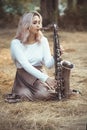 Portrait of a beautiful young woman sitting on the dry grass leanher head on saxophone, romantic blonde girl close eyes and Royalty Free Stock Photo