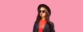 Portrait of beautiful young woman wearing black rock leather jacket, sunglasses, round hat on pink studio background Royalty Free Stock Photo