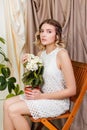 Portrait of a beautiful young woman in a white dress with a pot of artificial white flowers in her hands sitting on a chair Royalty Free Stock Photo