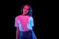 Portrait of beautiful young woman in white t-shirt and jeans posing against black studio background in neon light Royalty Free Stock Photo