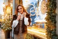 Portrait of a beautiful young woman who poses on the street near the elegantly decorated Christmas window, festive mood Royalty Free Stock Photo