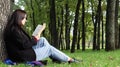 Portrait of a beautiful young woman who is sitting under a tree and reading her favorite book in a city park on green grass on a Royalty Free Stock Photo