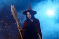 Portrait of beautiful young woman in witch halloween costume wear witches hat holding broom over spooky dark magic background Royalty Free Stock Photo