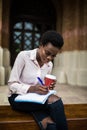 Portrait of beautiful young afro american student woman sitting outside on the bench writing in book and smiling, drinking coffee Royalty Free Stock Photo