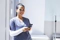 Portrait of biracial female doctor wearing scrubs using smartphon in hospital, copy space