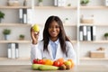 Portrait of black female dietologist holding apple, recommending fresh fruits and vegetables, sitting in medical office Royalty Free Stock Photo