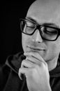 Portrait in black and white of a young bald man in black eyeglasses holding his chin with his hand while thinking looking down Royalty Free Stock Photo