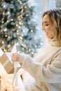 Portrait of blond hair woman in hoodie open present on Chrisrmas time. Elegant woman stand by big Christmas tree in Royalty Free Stock Photo