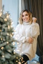 Portrait of blond hair woman in hoodie open present on Chrisrmas time. Elegant woman stand by big Christmas tree in Royalty Free Stock Photo