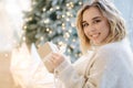 Portrait of blond hair woman in hoodie open present on Chrisrmas time. Elegant woman stand by big Christmas tree in Royalty Free Stock Photo