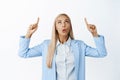 Portrait of blond saleswoman, pointing and looking up with amazed face expression, standing in blue suit over white