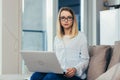 Portrait of a blonde woman looking at the camera sitting at home on the couch and holding a laptop Royalty Free Stock Photo