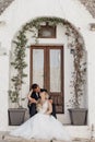 Portrait of blonde bride in white dress and brunet groom in suit kissing and sitting near old city house with arch from Royalty Free Stock Photo