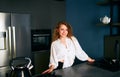 Portrait of blonde caucasian woman standing and smiling in her kitchen leaning on the table. Young cheerful adult female Royalty Free Stock Photo