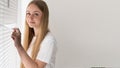 A portrait of a blonde girl standing by the window looks behind the blinds to the left of the space for the text. White Royalty Free Stock Photo