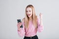 Portrait of a blonde girl who holds a smartphone and shows the gesture of the winner in the studio on a white background. Royalty Free Stock Photo