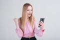Portrait of a blonde girl who holds a smartphone and shows the gesture of the winner in the studio on a white background. Royalty Free Stock Photo