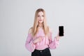 Portrait of a blonde girl who shows the smartphone screen and points her finger at it in the studio on a white background. Royalty Free Stock Photo