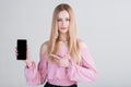 Portrait of a blonde girl who shows the smartphone screen and points her finger at it in the studio on a white background. Royalty Free Stock Photo