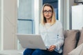 Portrait of a blonde woman looking at the camera sitting at home on the couch and holding a laptop Royalty Free Stock Photo