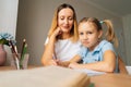 Portrait of blonde primary daughter doing homework with young mother sitting at home table by window, looking at camera. Royalty Free Stock Photo