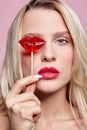 Portrait of blonde woman closing by candy her eye.. Red female lips shape lollipop. Sweet tooth concept Royalty Free Stock Photo