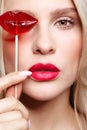 Portrait of blonde woman closing by candy her eye.. Red female lips shape lollipop. Sweet tooth concept Royalty Free Stock Photo