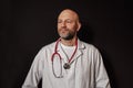 Portrait of a bold slim doctor on a dark background. Caucasian male in 40s, grey beard, looking away from camera, wearing white co Royalty Free Stock Photo