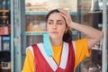 Portrait of a bored employee in uniform, with a medical mask over his ear. Showcase with products in the background. Concept of