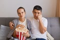 Portrait of bored sad upset wife and husband sitting on sofa with pop-corn and remote control, watching boring movie, looking at Royalty Free Stock Photo