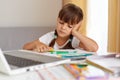 Portrait of bored schoolgirl wearing white t shirt, sitting at table, in front of notebook, having sad facial expression, being Royalty Free Stock Photo