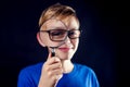 A portrait of a boy with eyeglasses looks through a magnifying glass in front of dark background. Children and education concept Royalty Free Stock Photo