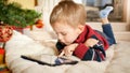 Portrait of little boy lying on blanket next to Christmas tree and playing on digital tablet computer Royalty Free Stock Photo