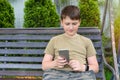 A portrait of a boy with a phone in his hands, sitting on the bench in the backyard. Texting in a cell phone. A teenager reading a Royalty Free Stock Photo